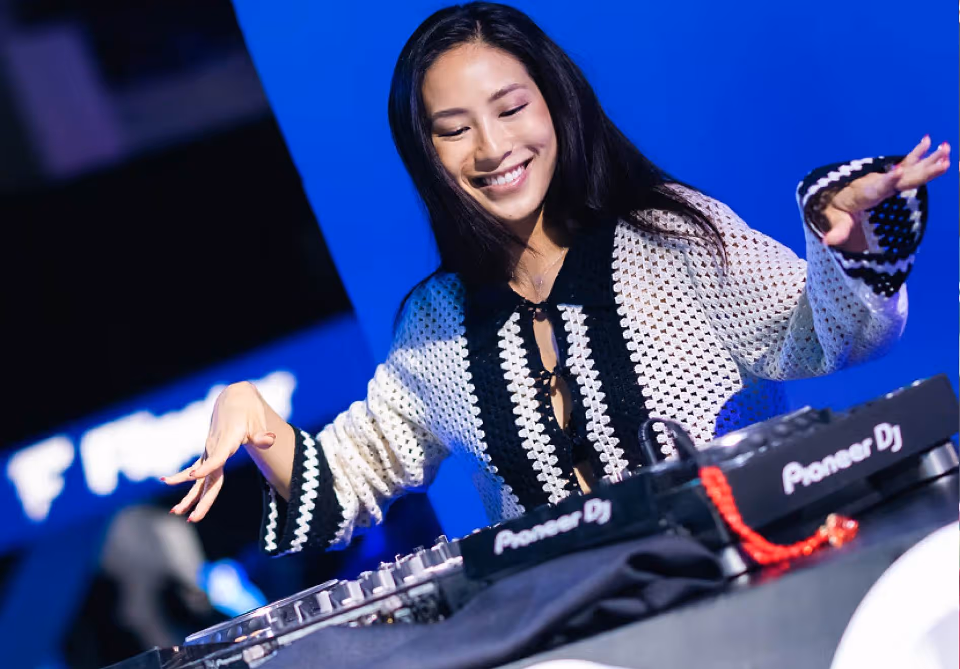 Smiling female DJ wearing a black and white crocheted sweater playing music on Pioneer DJ equipment against a blue background.